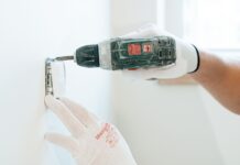 Ręce w rękawicach montują wiertarką gniazdko elektryczne w ścianie