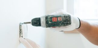 Ręce w rękawicach montują wiertarką gniazdko elektryczne w ścianie