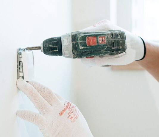 Ręce w rękawicach montują wiertarką gniazdko elektryczne w ścianie