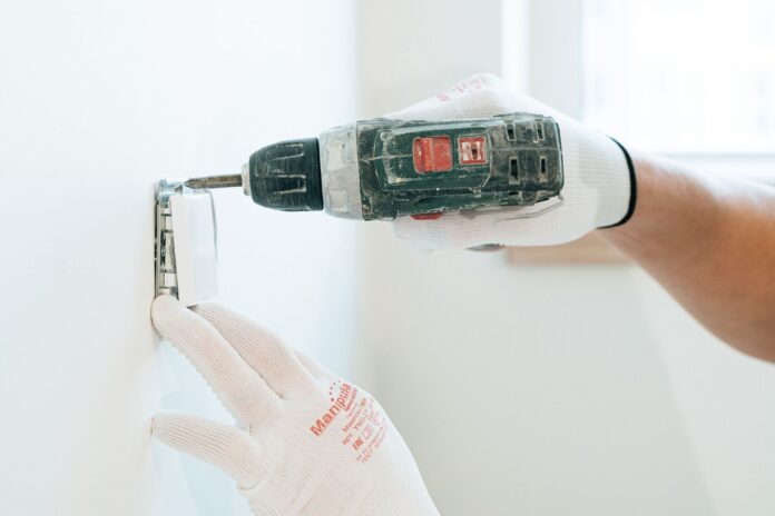 Ręce w rękawicach montują wiertarką gniazdko elektryczne w ścianie